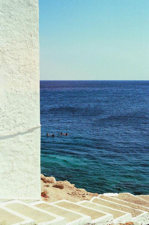 Three people swim in the ocean next to a white wall and stairs leading down to the water.