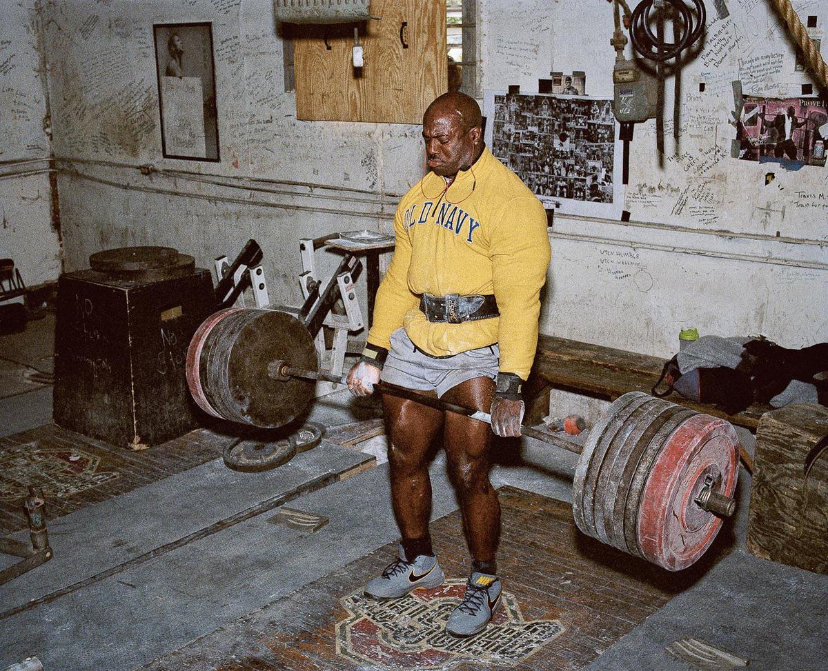A man in a yellow sweatshirt and gray shorts lifts a barbell in a gym.