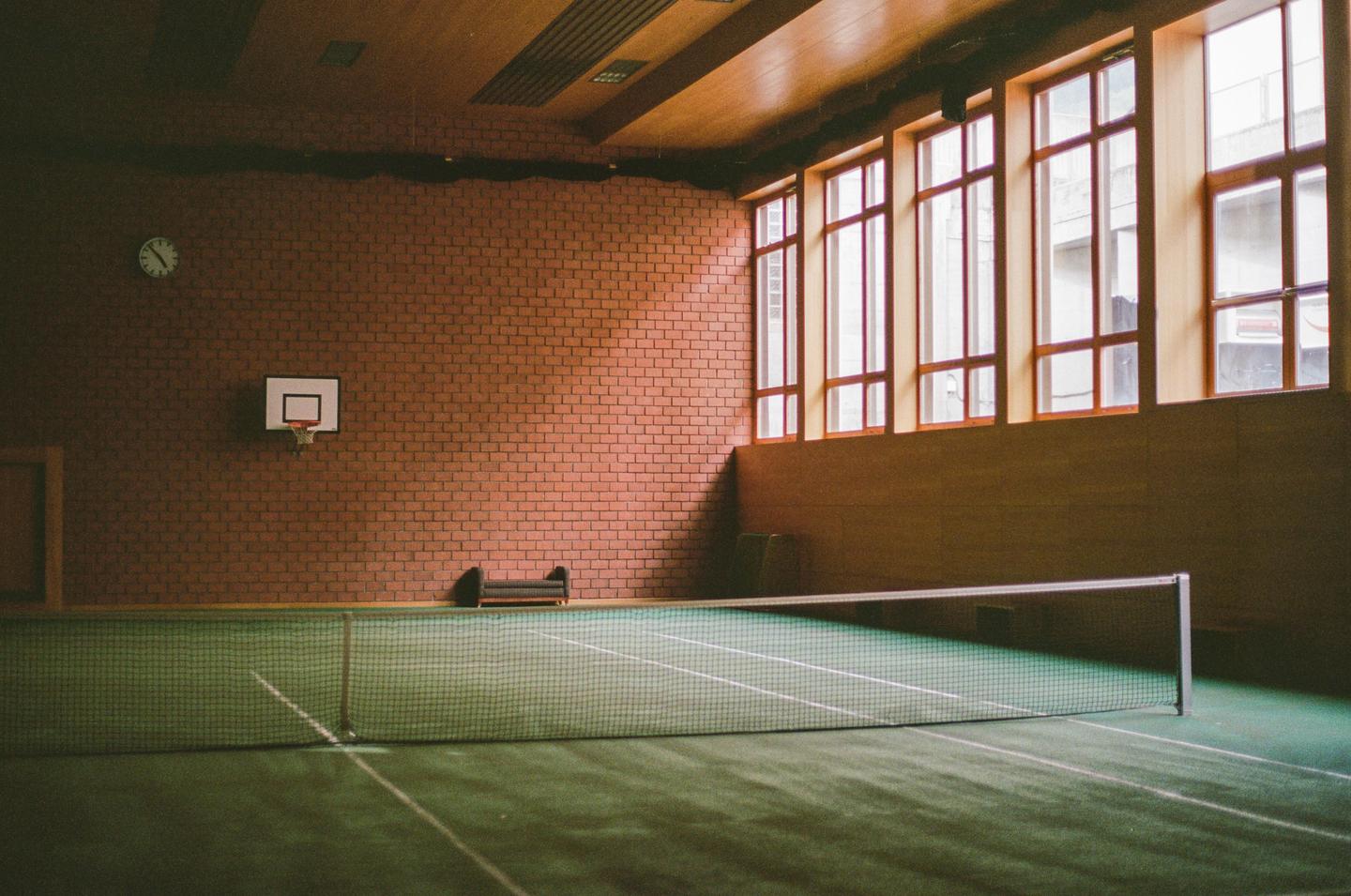 A tennis court with a net and a basketball hoop is visible inside a gymnasium with brick walls and large windows.