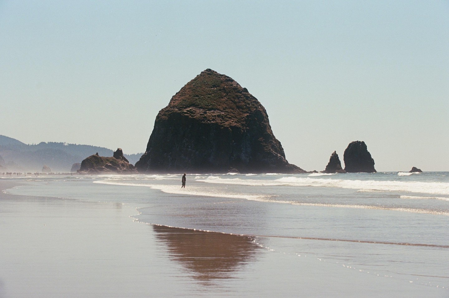A person stands in the shallow water of a beach with large rock formations in the background.