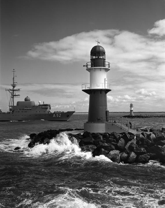 A large ship passes a lighthouse on a rocky jetty with waves crashing against it.