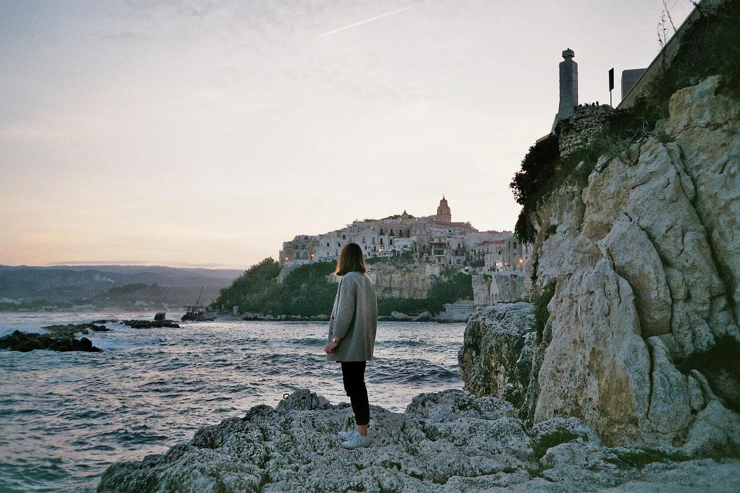 A woman stands on a rocky shore looking out at a town built on a cliff overlooking the sea at dusk.
