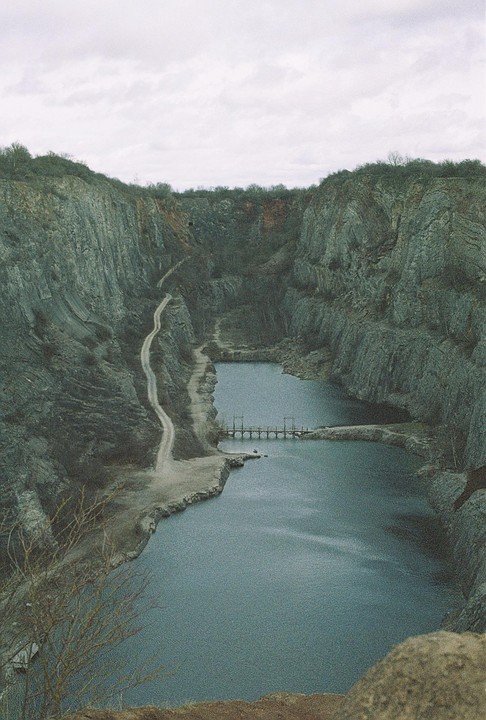 A winding dirt road leads down into a quarry filled with water, with a small bridge crossing the water.
