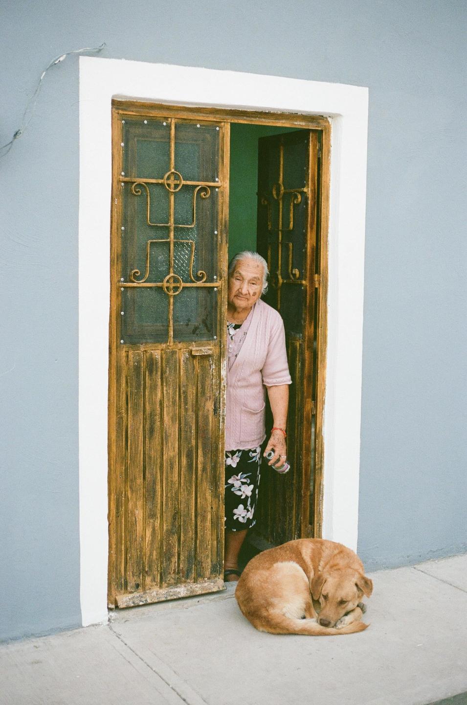 An elderly woman peeks out from behind a wooden door next to a sleeping dog.