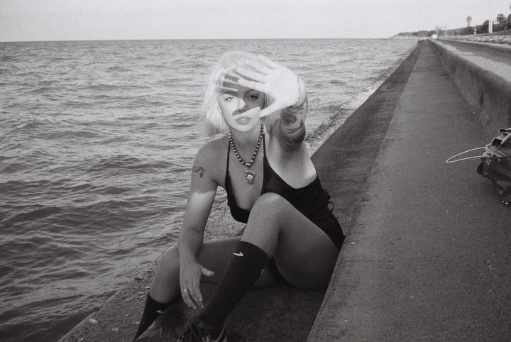 A woman with blonde hair sits on a concrete ledge next to the ocean, holding her hand up to block the sun.
