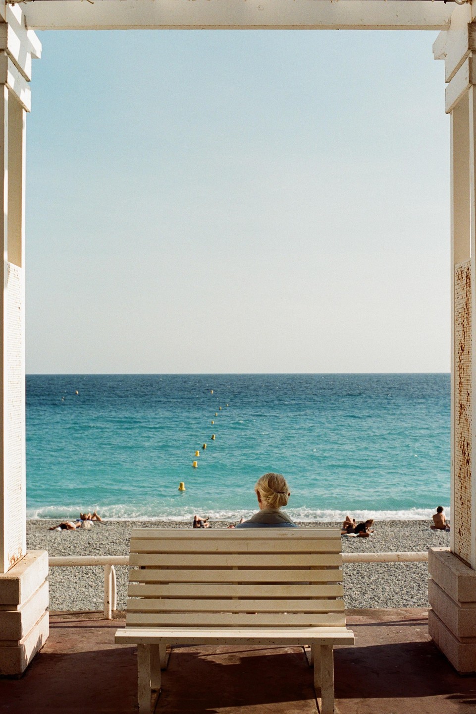 A woman sits on a bench looking out at the ocean from a covered promenade.
