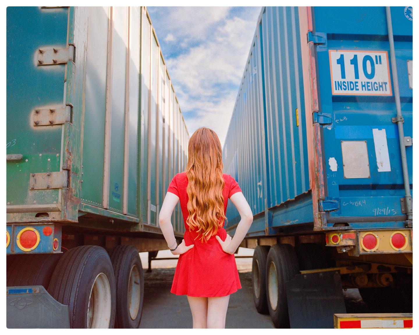 A woman with long red hair stands between two large shipping containers under a bright sky.
