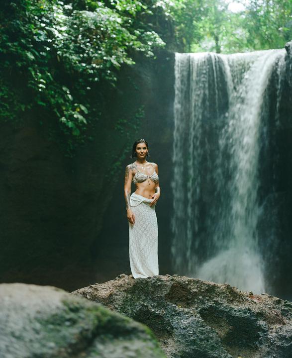 A woman stands on a rock in front of a waterfall in a lush, green environment.