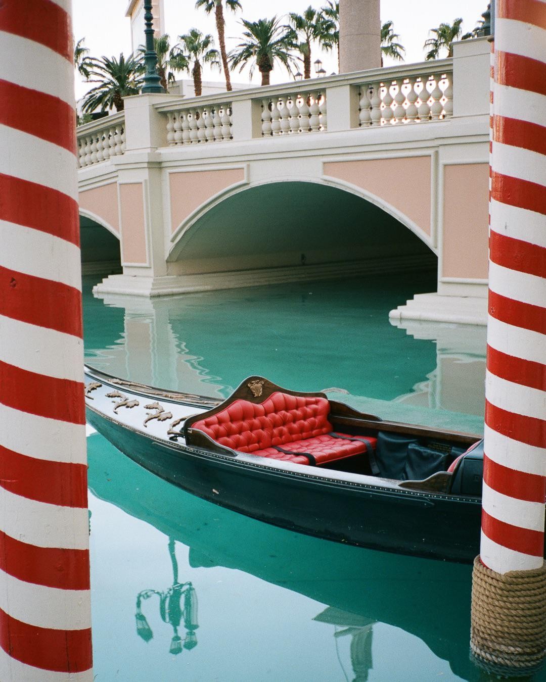 A gondola with red seats floats on turquoise water next to a bridge with palm trees in the background.
