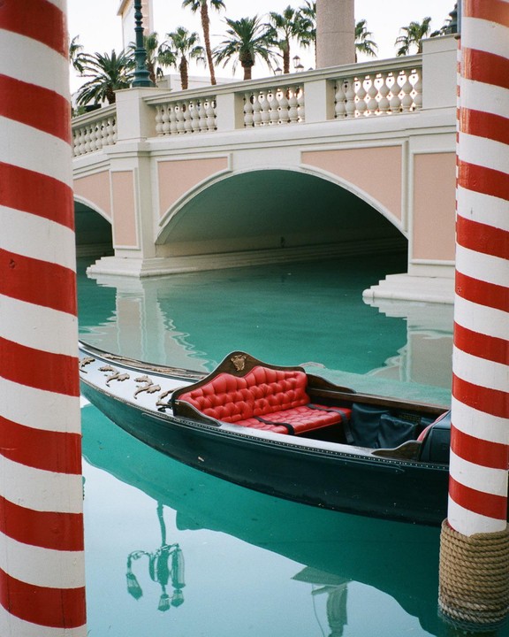 A gondola with red seats floats on turquoise water next to a bridge with palm trees in the background.