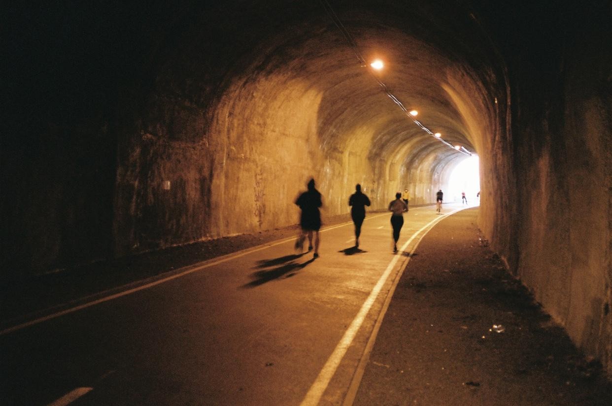 Three people run through a tunnel with lights on the ceiling and a bright light at the end.