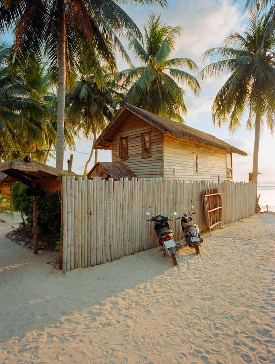 A wooden house with a thatched roof stands on a sandy beach next to a bamboo fence, with two motorbikes parked nearby and palm trees overhead.