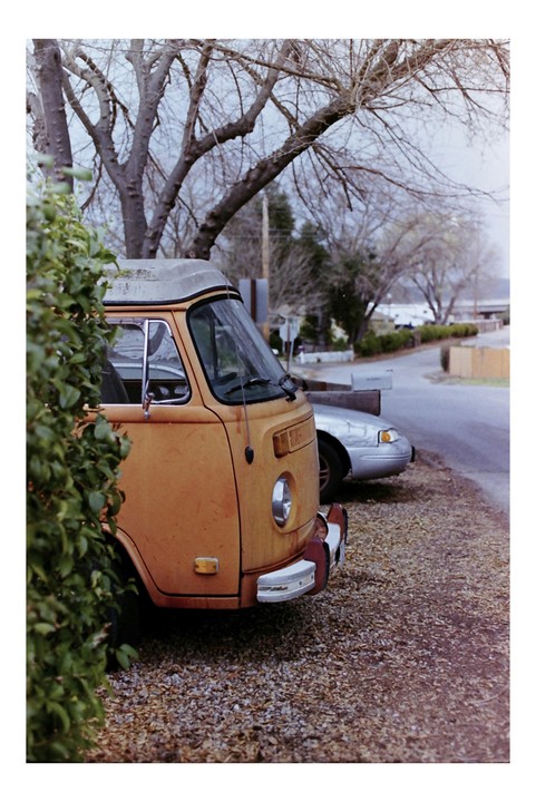 An orange van is parked on a gravel driveway next to a green bush, with a street and bare trees in the background.
