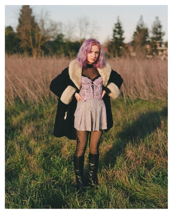 A woman with purple hair stands in a field wearing a black coat with fur trim, a pink corset, a skirt, fishnet stockings, and boots.