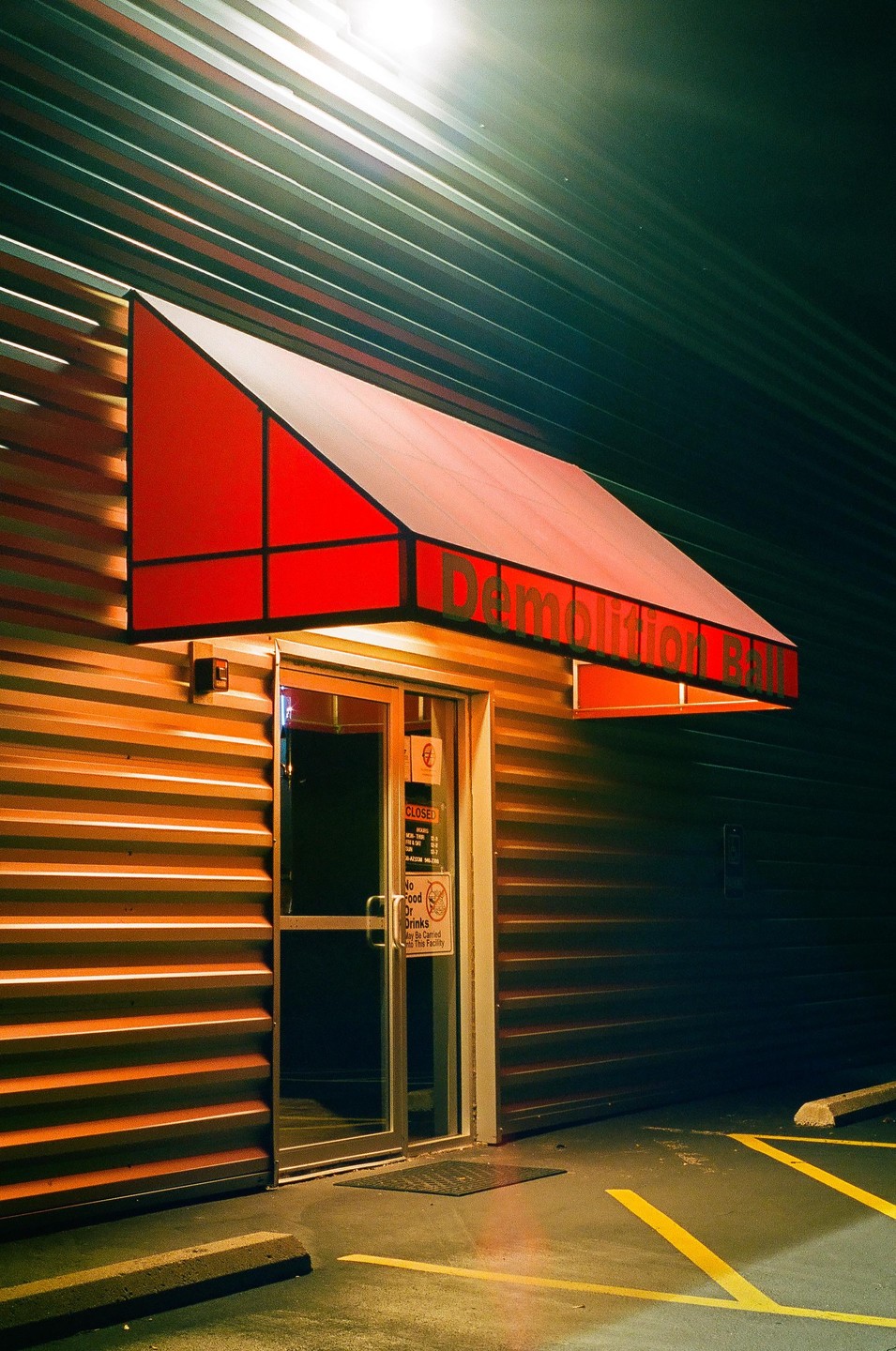A red awning with "Demolition Bill" written on it hangs over a glass door on a corrugated metal building at night.