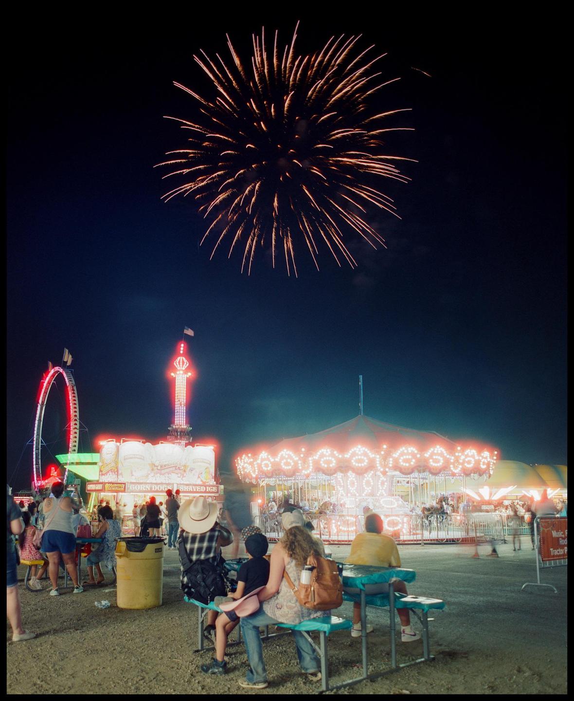 A firework explodes over a carnival with people sitting on benches in the foreground.