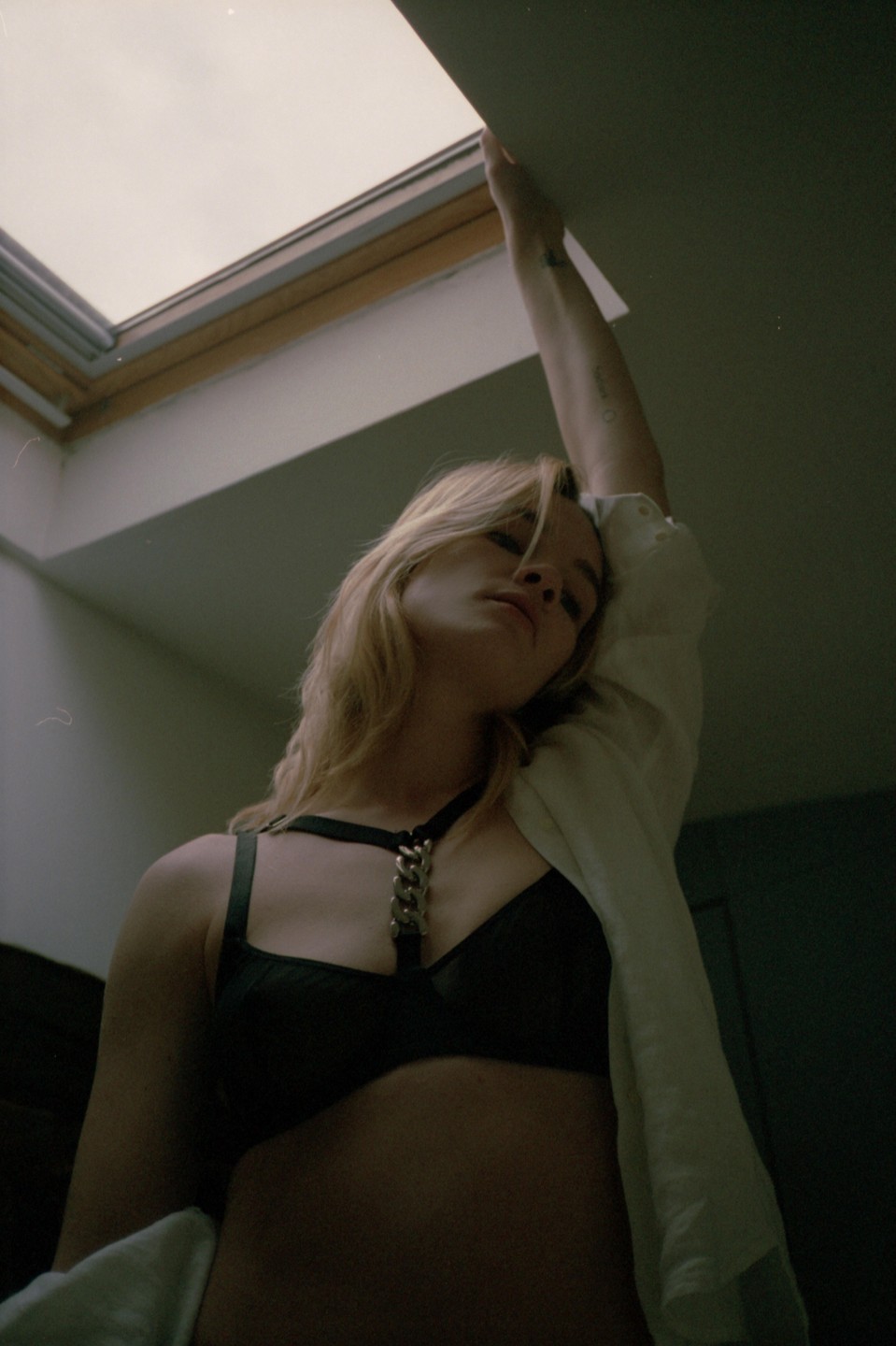 A woman in a black bra with a chain detail and an open white shirt stretches her arm towards a skylight.