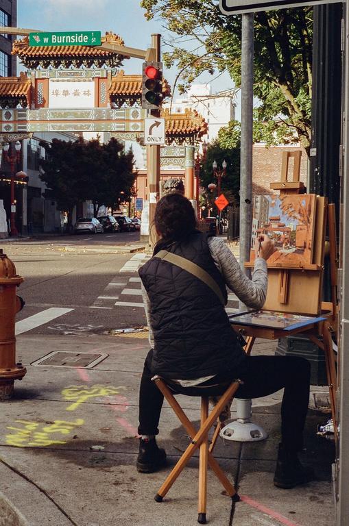 A woman sits on a folding stool painting a street scene with a red traffic light and an ornate gate in the background.