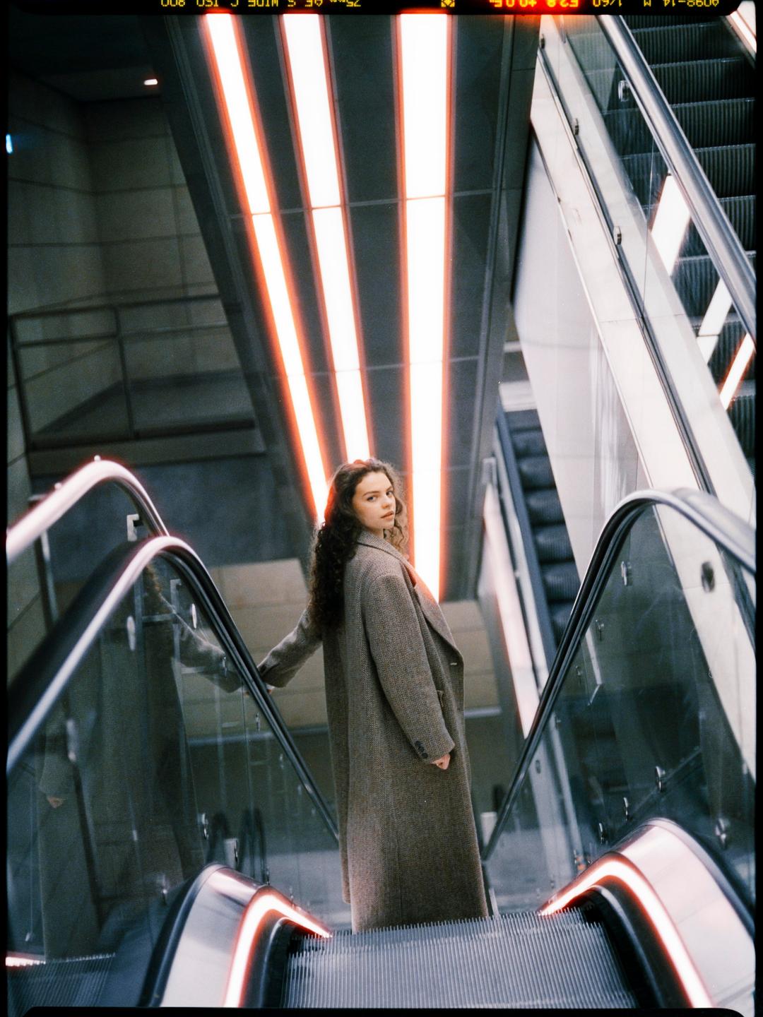 A woman in a long coat stands on an escalator looking back at the viewer.