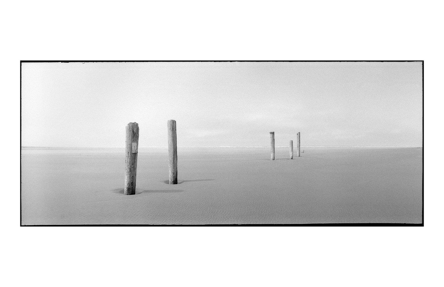 A series of wooden posts stands in the sand on a wide, flat beach under a bright sky.