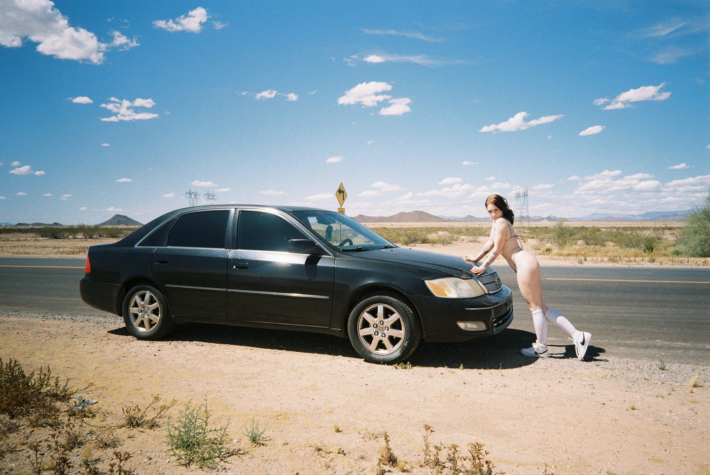 A woman in a bodysuit leans on the hood of a black car on the side of a desert road under a blue sky with clouds.