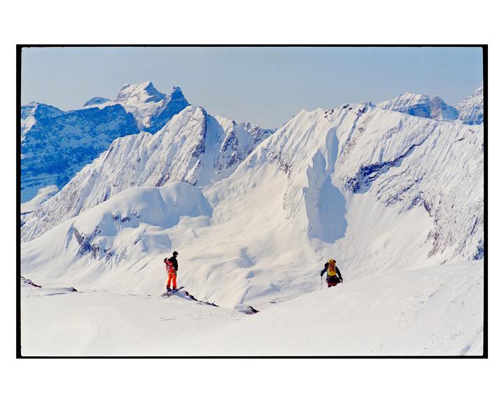 Two people are in a snowy mountain landscape, one on a snowboard and the other walking with poles.