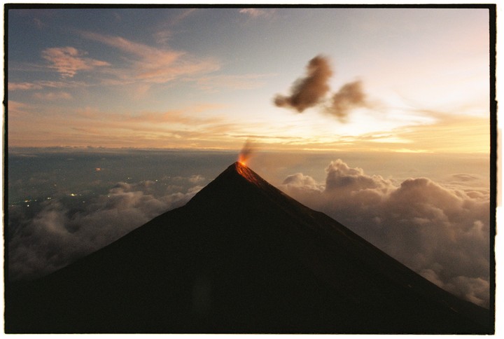 A volcano erupts with glowing lava and smoke against a sunset sky with clouds below.