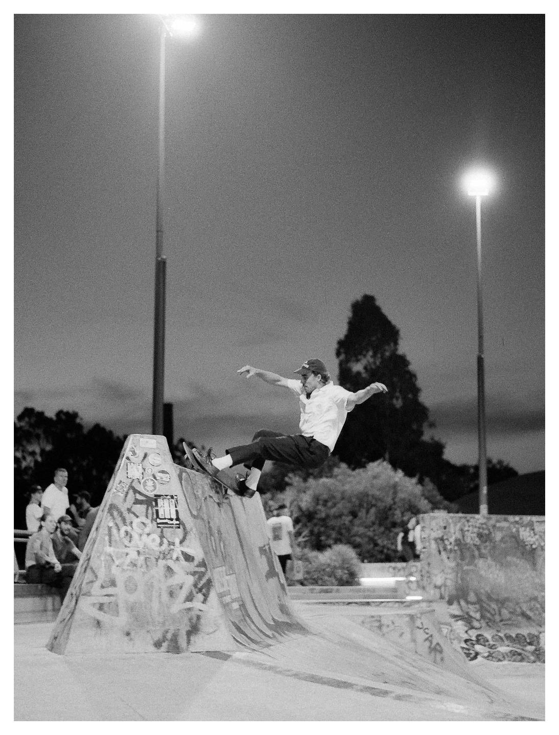 A man on a skateboard is airborne over a graffiti-covered ramp at a skatepark at night.