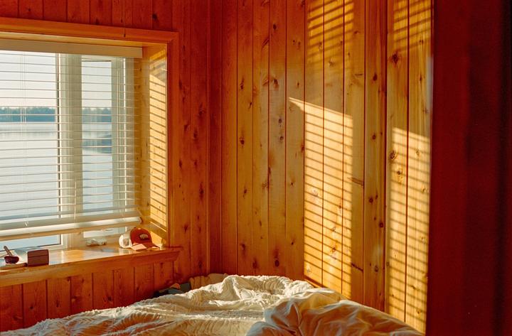 Sunlight streams through window blinds onto a wooden wall and an unmade bed.