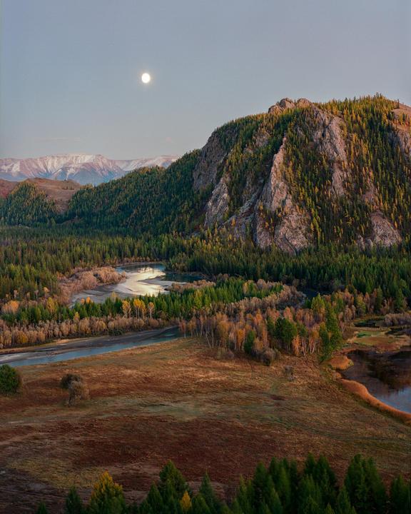 A full moon hangs over a winding river and a forest-covered mountain with snow-capped peaks in the distance.