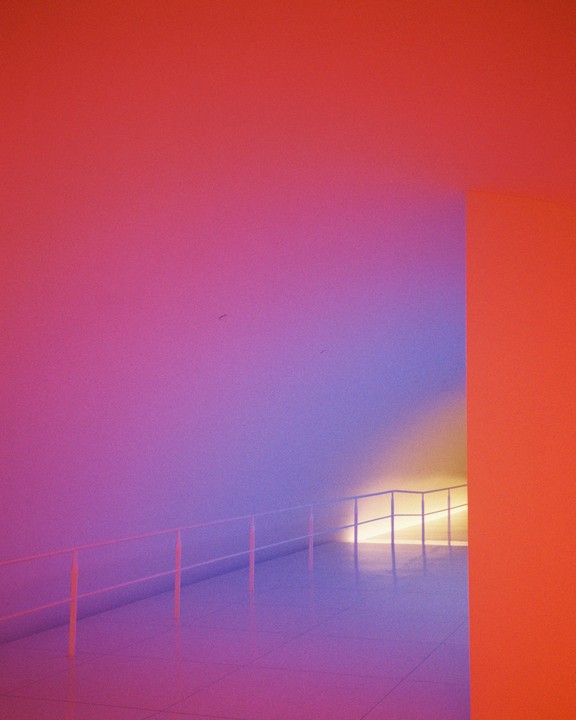 A brightly lit tunnel with a railing is illuminated by a gradient of colors.