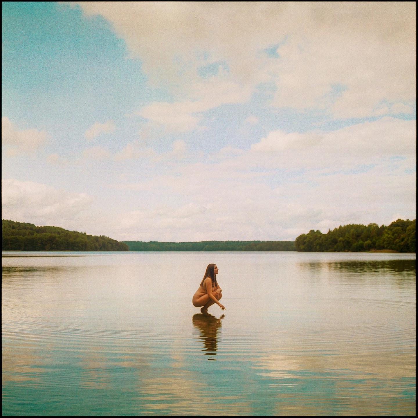 A woman crouches in a lake with trees on the far bank under a cloudy sky.