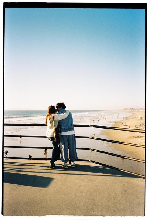 A couple stands on a pier, looking out at the ocean and beach.