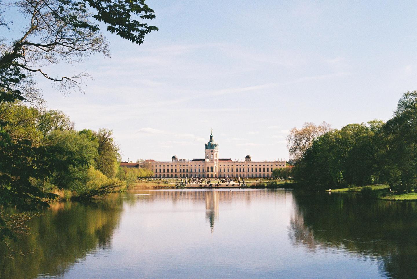 A large palace with a prominent central dome is reflected in the calm water of a lake, surrounded by lush green trees under a clear sky.
