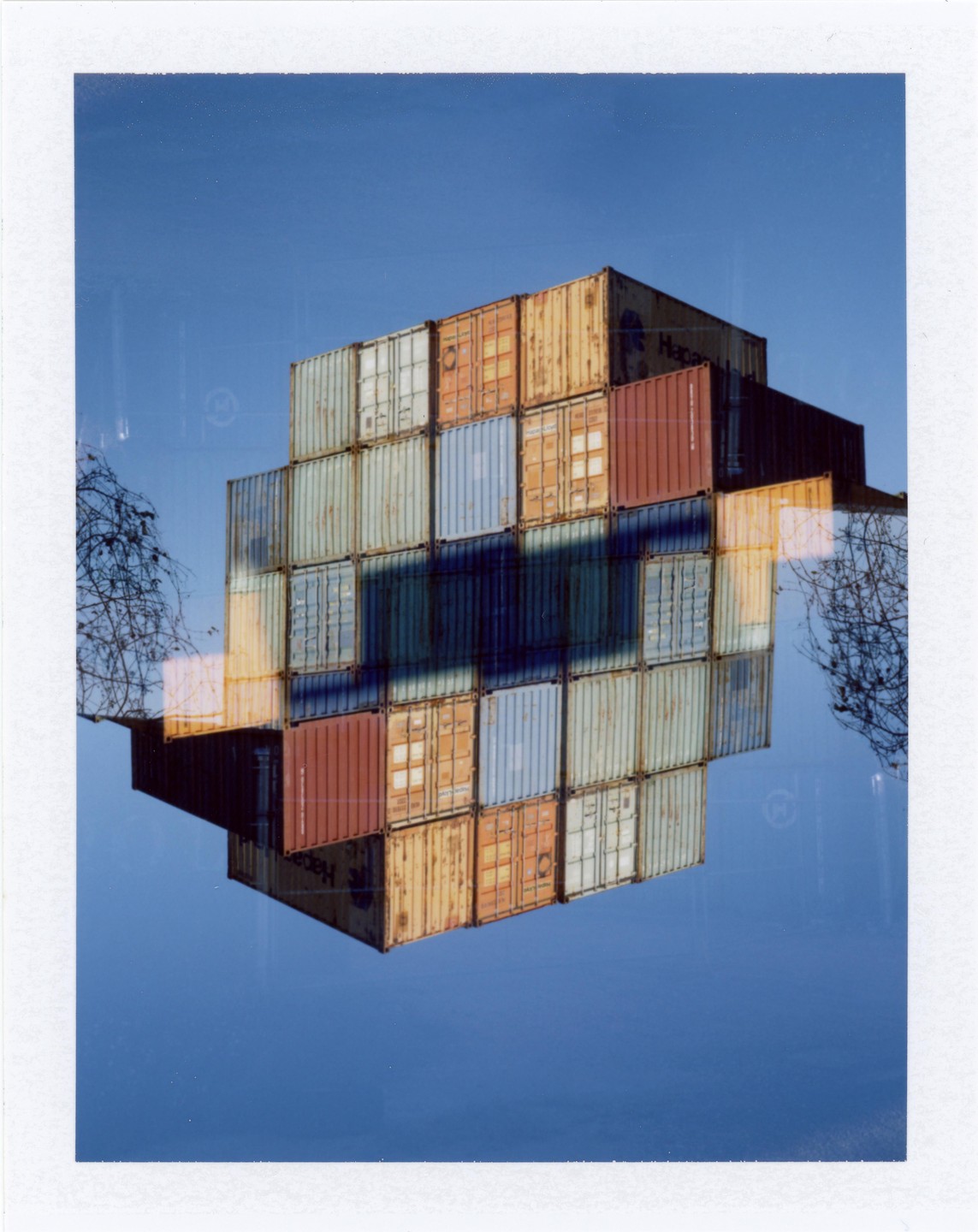 A double exposure shows a stack of colorful shipping containers against a blue sky, with tree branches visible on the sides.