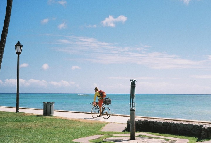 A person rides a bicycle along a paved path next to the ocean under a blue sky with scattered clouds.