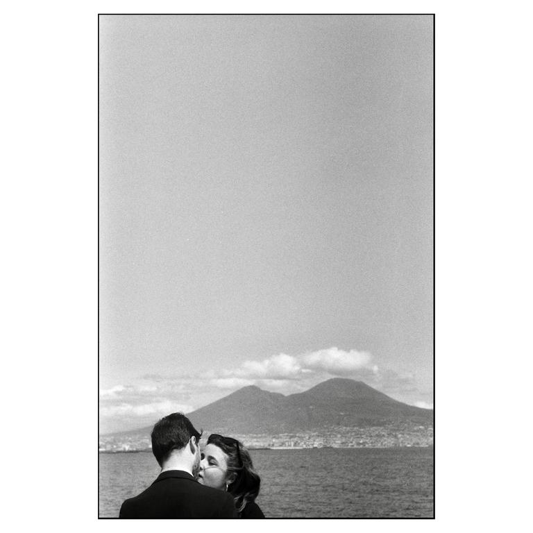 A couple stands in front of a body of water with mountains and a city in the background.