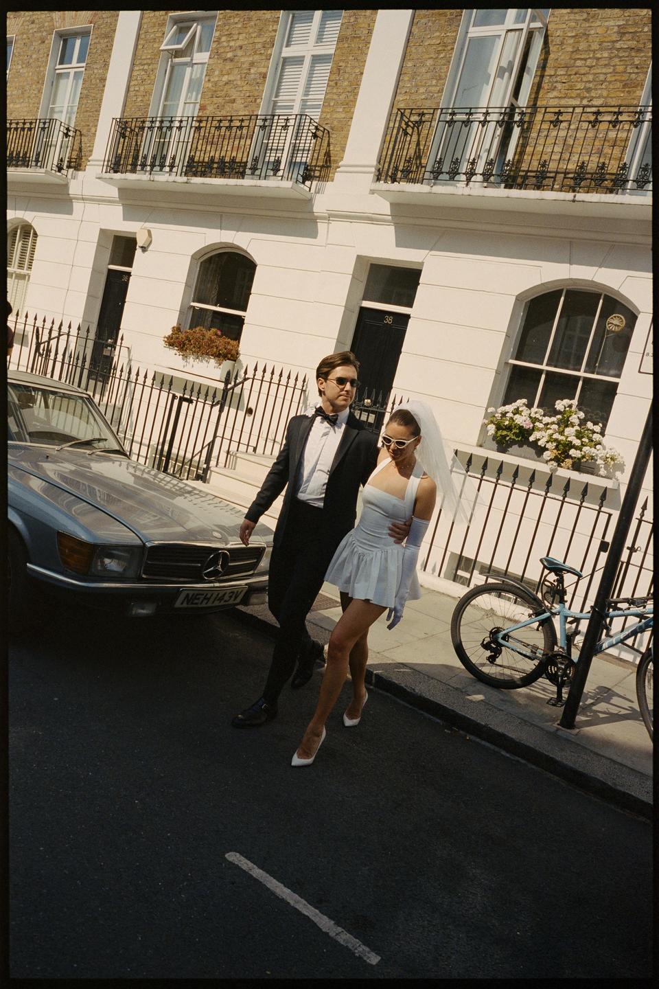 A couple in wedding attire walks along a city street next to a parked car.