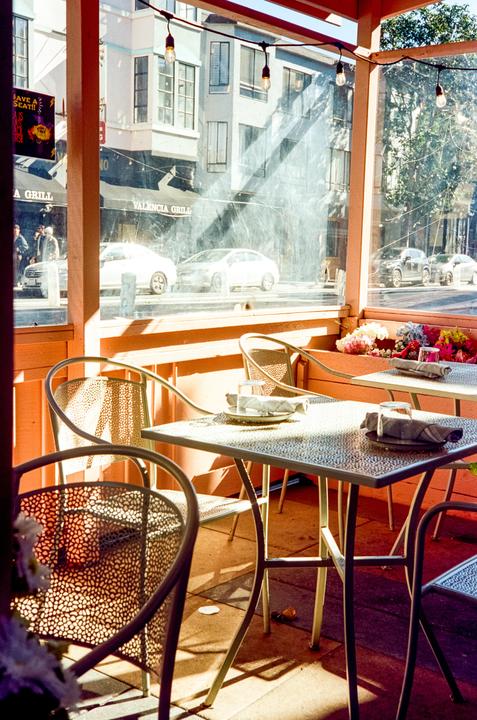 Sunlight streams into an outdoor dining area with tables and chairs set for two, overlooking a street with cars and buildings.
