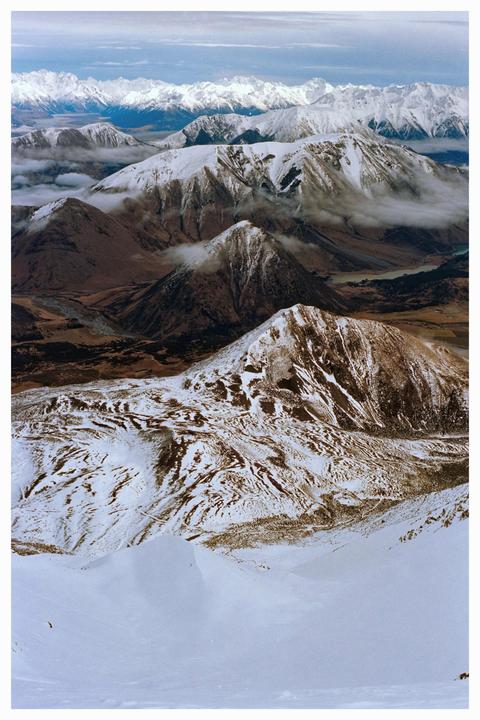 An aerial view shows a vast mountain range with snow-capped peaks and valleys partially obscured by clouds.
