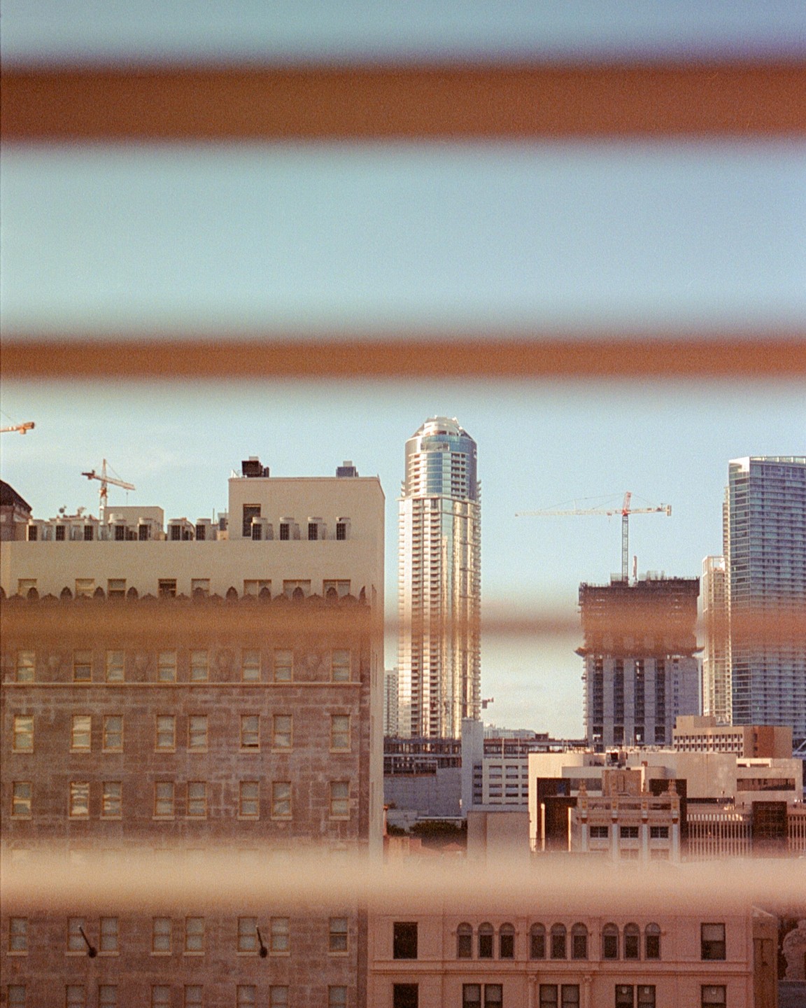 A cityscape with several tall buildings and construction cranes is viewed through horizontal blinds.