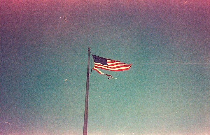 A tattered American flag flies from a flagpole against a colorful sky.