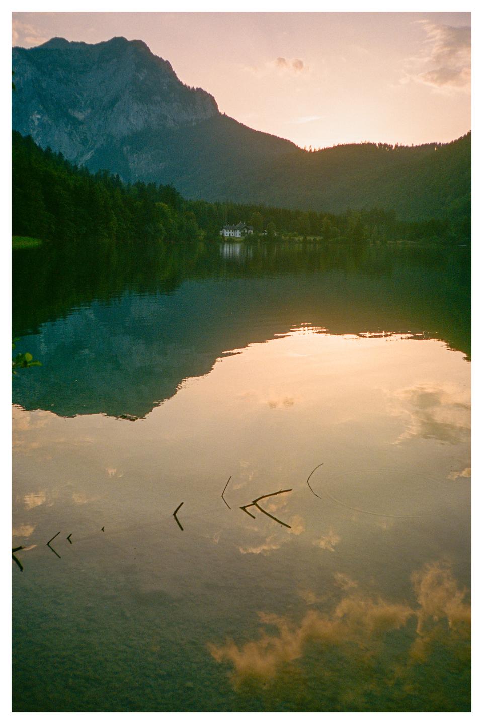 A large mountain and forest reflect in a lake with a house on the far shore under a sunset sky.