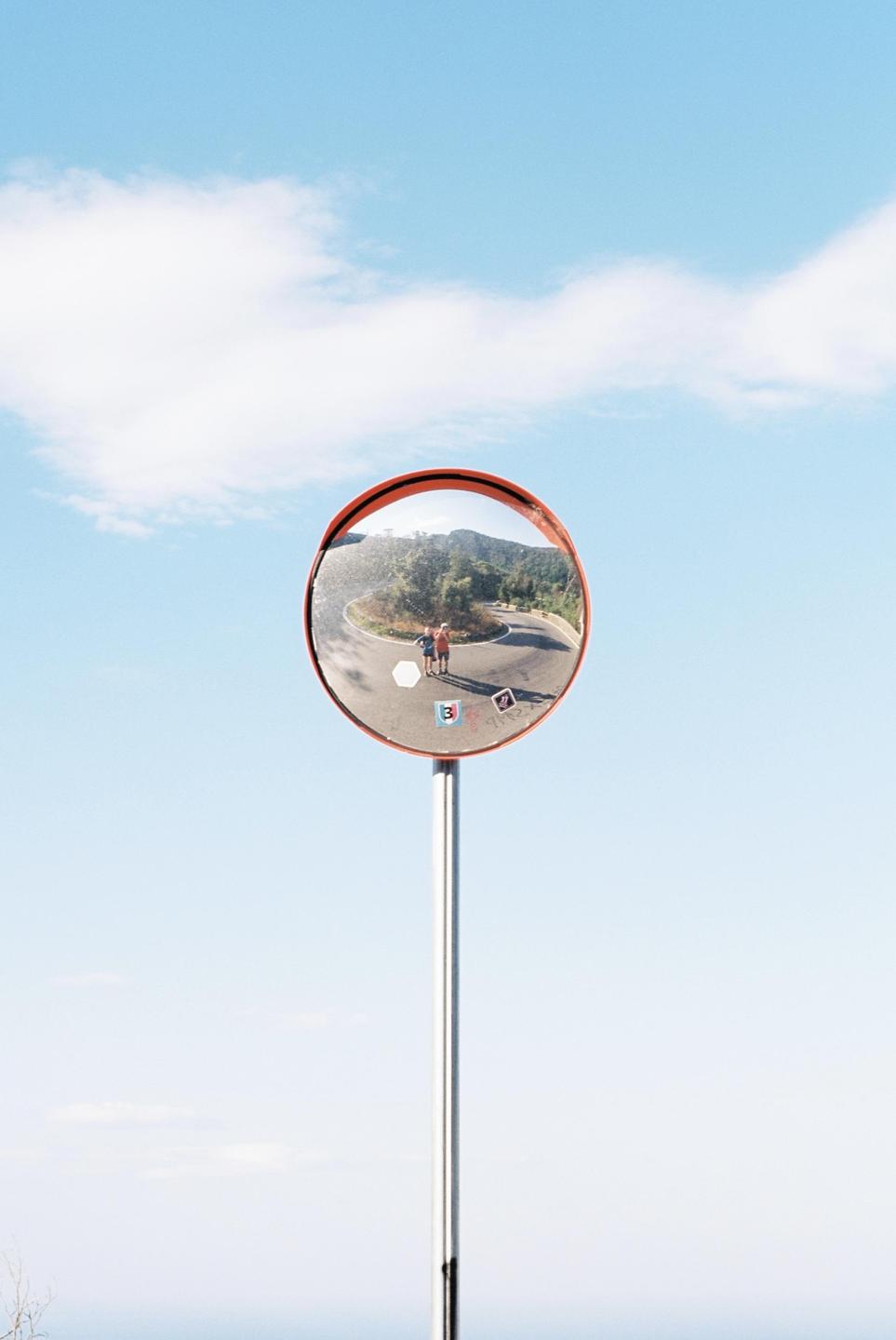 A convex mirror on a pole reflects a winding road with two people and mountains under a blue sky.