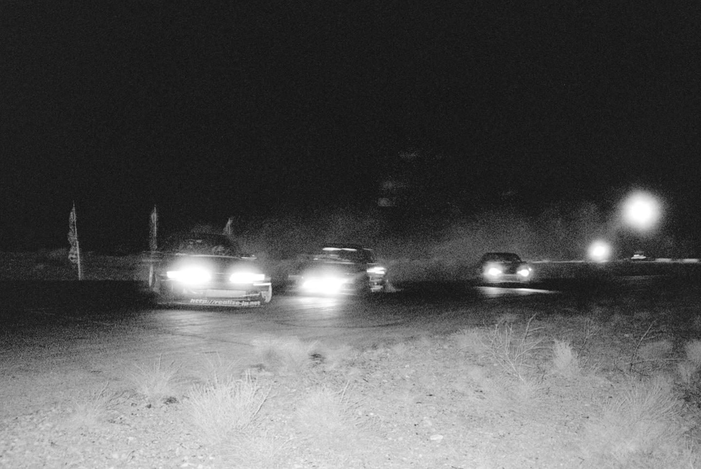 Three cars with bright headlights are driving on a dirt track at night, with some flags visible on the left.