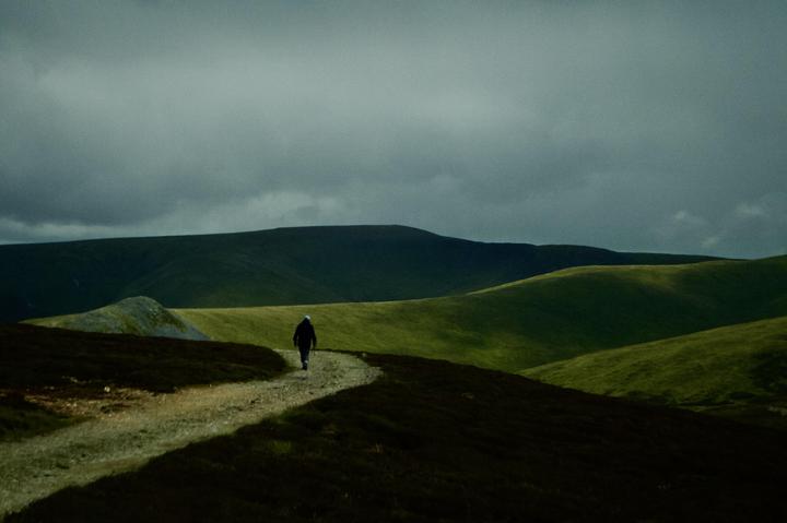 A person walks along a winding path through rolling hills under a cloudy sky.
