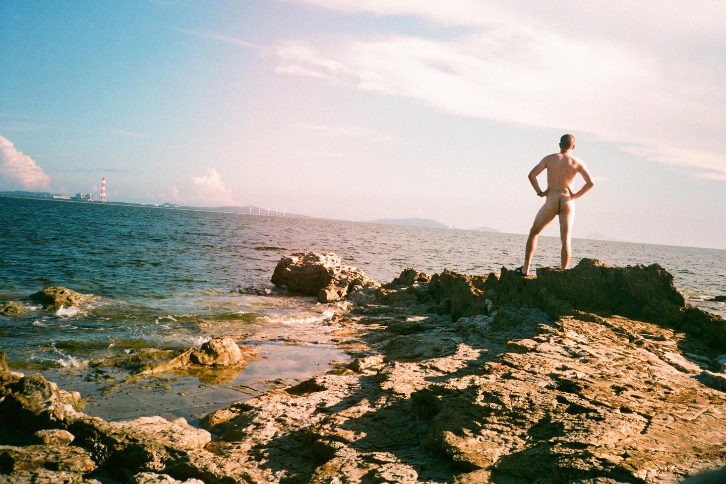 A man stands naked on a rocky shore looking out at the ocean with a lighthouse and wind turbines in the distance.