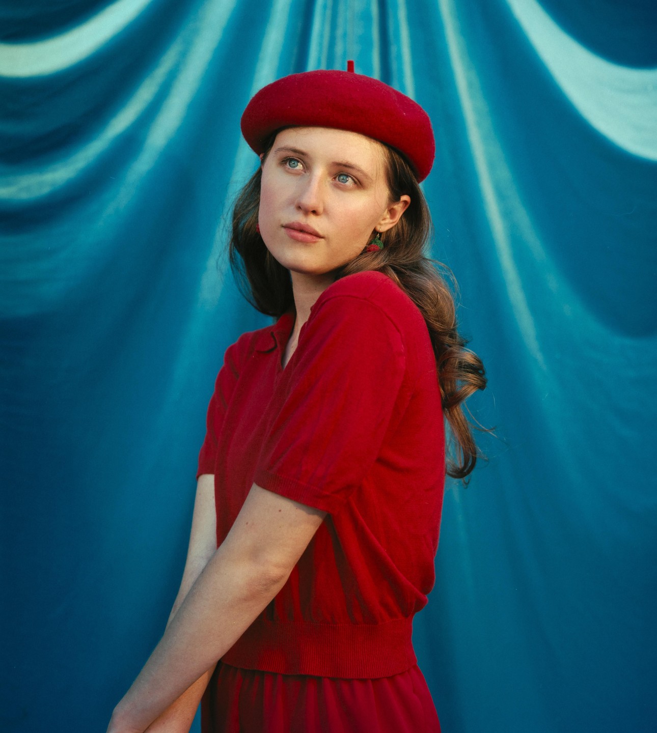 A woman in a red beret and red shirt looks up against a blue draped background.