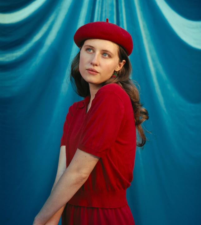 A woman in a red beret and red shirt looks up against a blue draped background.