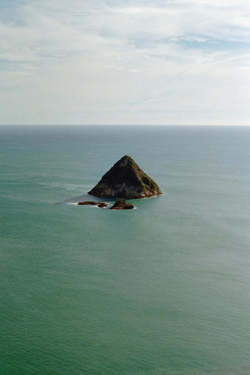 A small, pointy island covered in green foliage rises from the ocean under a cloudy sky.